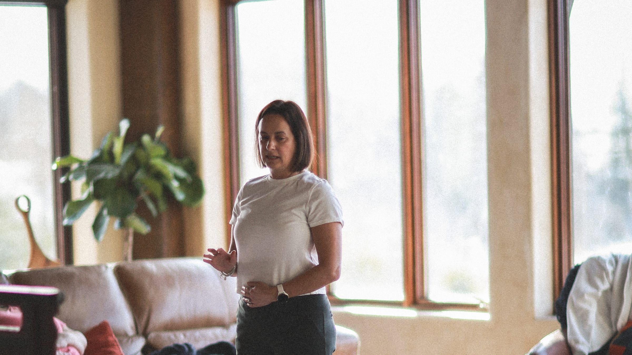 Louise demonstrating diaphragmatic breathing with a hand on her belly, in a bright window-lined room