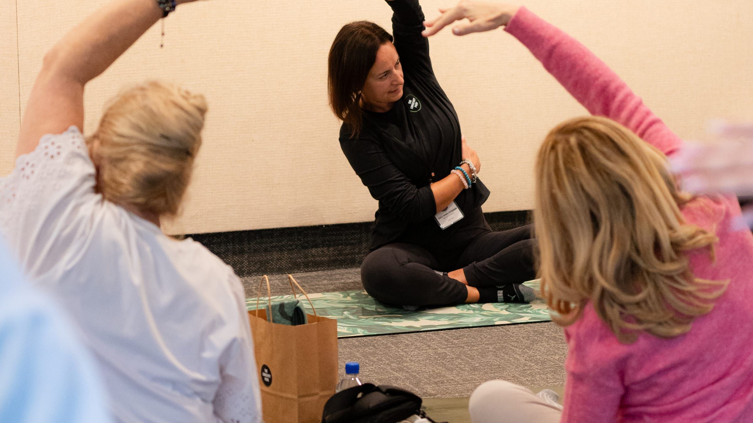 Louise seated on a mat leading a group through a gentle side stretch during a workplace wellness session
